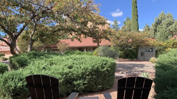 Two Adirondack chairs facing a gravel path with a small table, surrounded by lush greenery and a building with a red-tiled roof in the background.