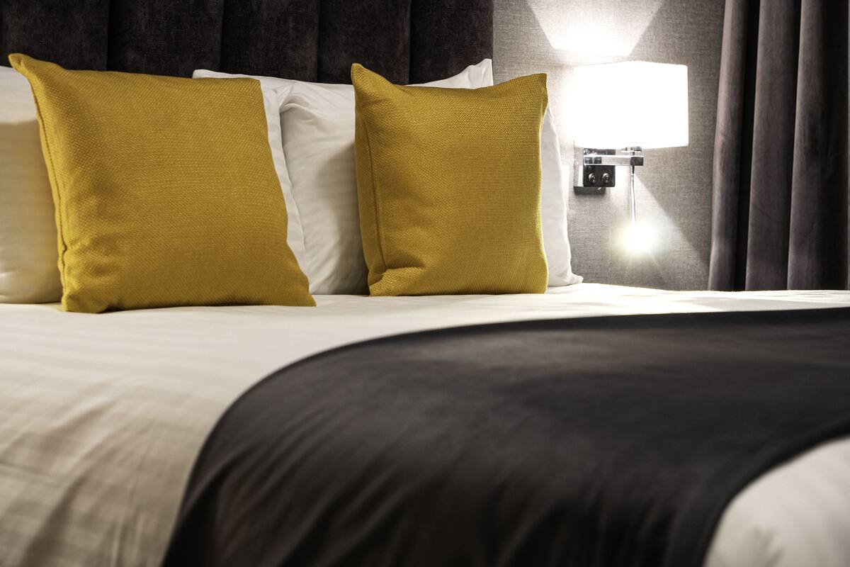 Een net opgemaakt bed met een zwart-wit dekbed, twee mosterdgele kussens en een wandlamp.
