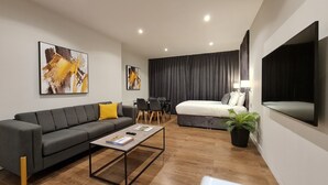 Een moderne hotelkamer met een grijze bank, een klein bijzettafeltje, een zithoek met stoelen, een bed met een zwart hoofdeinde en een grote spiegel.