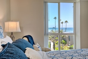 Quarto com uma janela ampla que oferece vista para a praia e palmeiras.