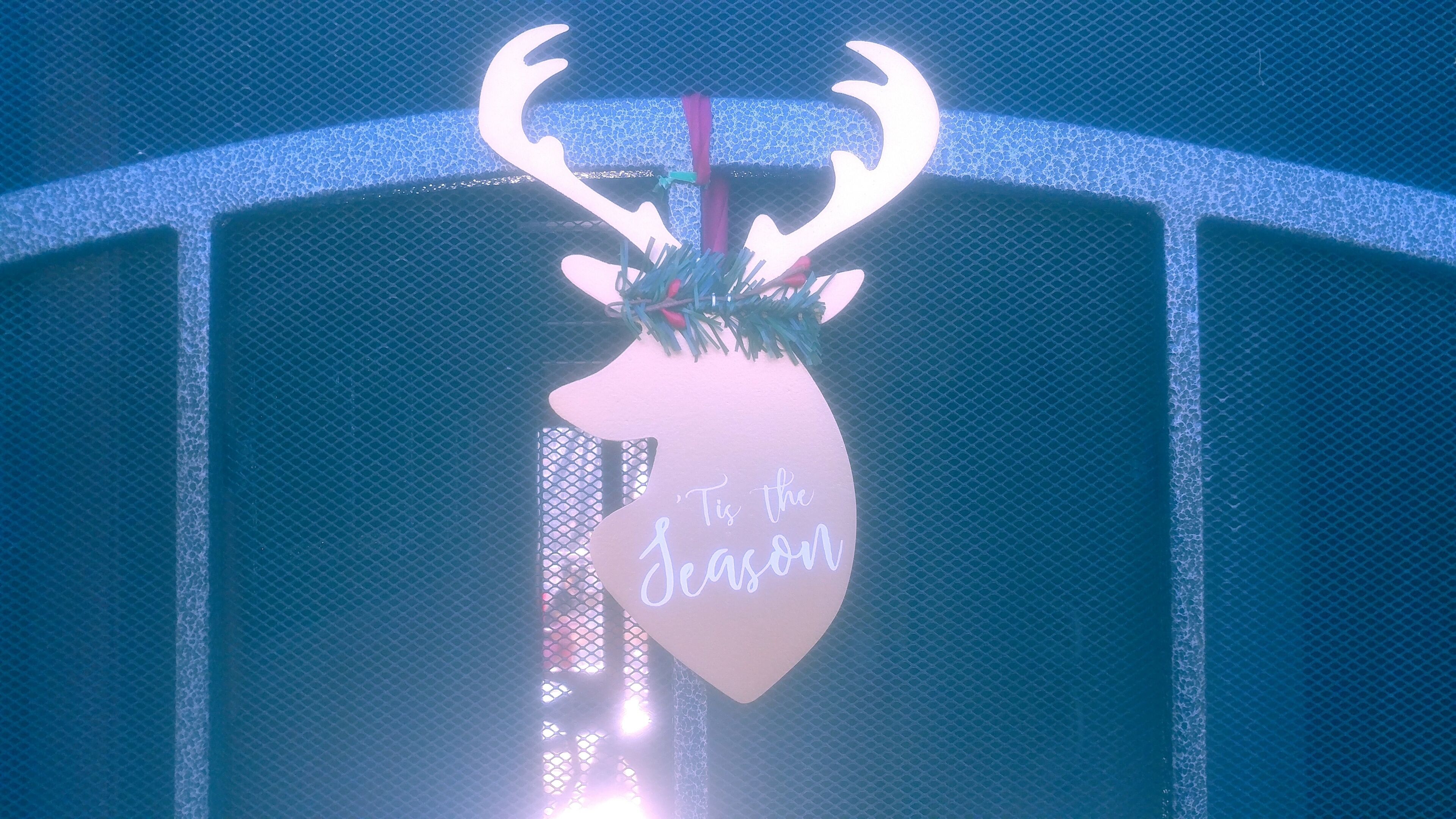 Una señal decorativa con forma de corazón, con la cabeza de un reno y el texto 'It's the Season' colgado sobre un fondo de malla azul.