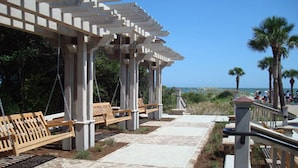 Un espace extérieur abrité, doté de bancs et d’une pergola, un sentier et des palmiers en arrière-plan.