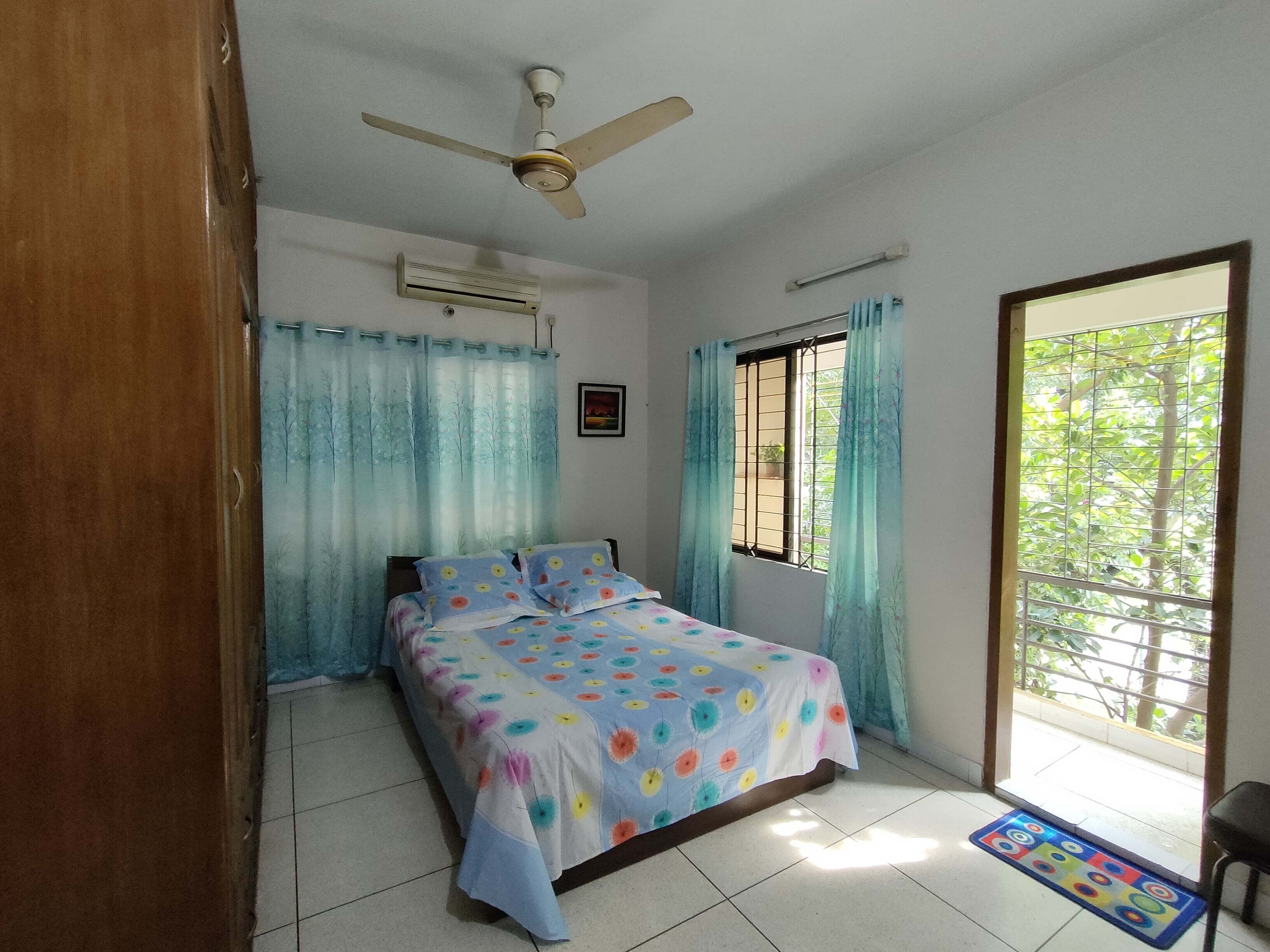 Un dormitorio con una cama, ventilador de techo, aire acondicionado, ventana con rejas y vista a la vegetación exterior.