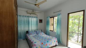 Un dormitorio con una cama, ventilador de techo, aire acondicionado, ventana con rejas y vista a la vegetación exterior.