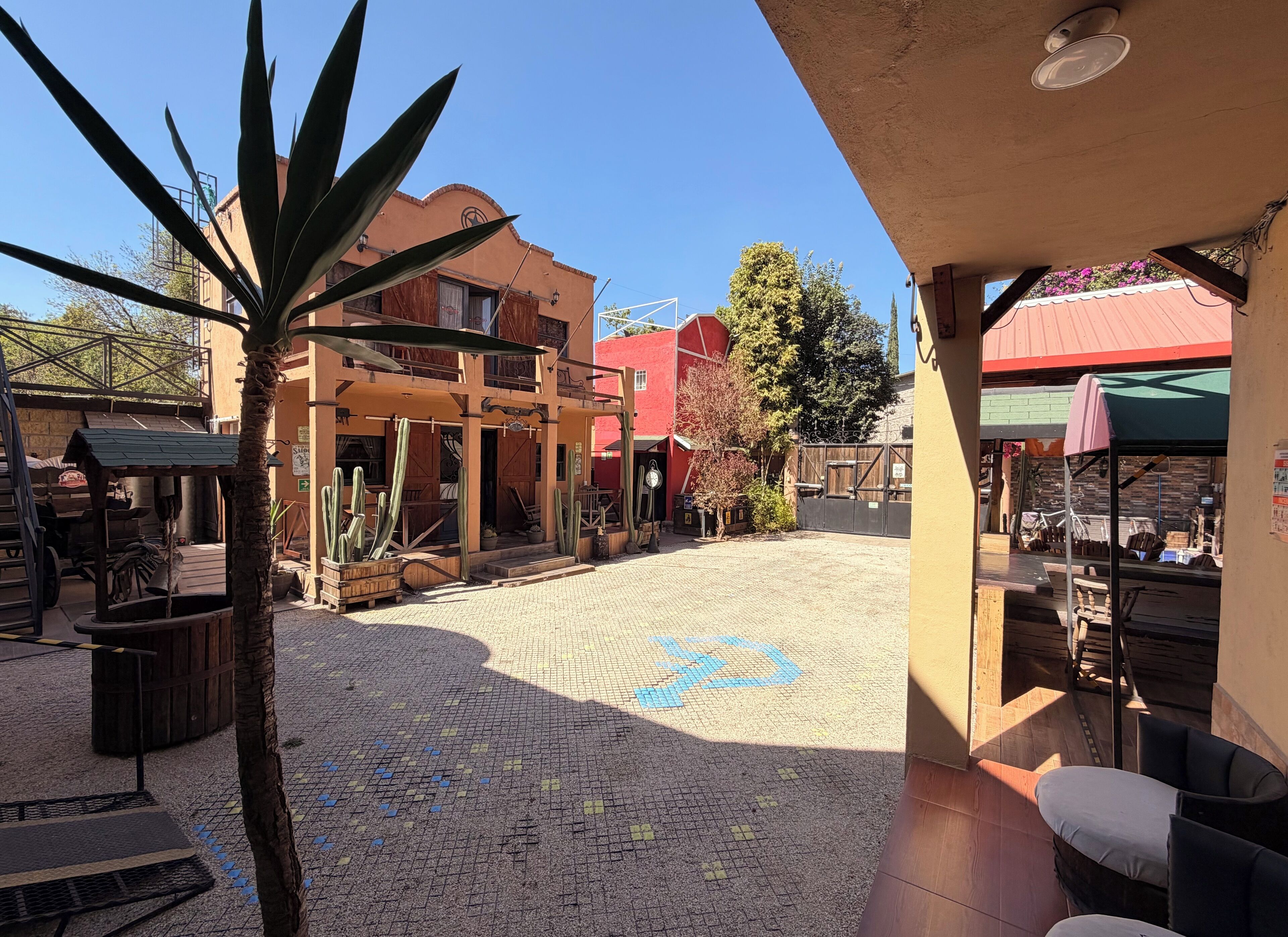 Un patio con suelo de baldosas, plantas de cactus, mobiliario de madera y un edificio de techo rojo.