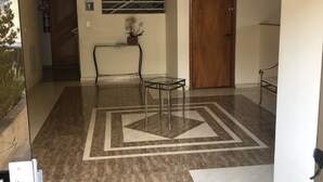 A spacious hallway with a patterned floor, a glass table with metal legs, and a wooden door. - Aconchego lar doce lar (Poços de Caldas)