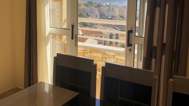 A room with a dining table and chairs, a window with curtains, and a view of buildings and mountains. - Aconchego lar doce lar (Poços de Caldas)