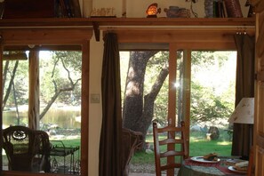 Una habitación con interior de madera, un ventanal con cortinas, una silla y una mesa con una lámpara.