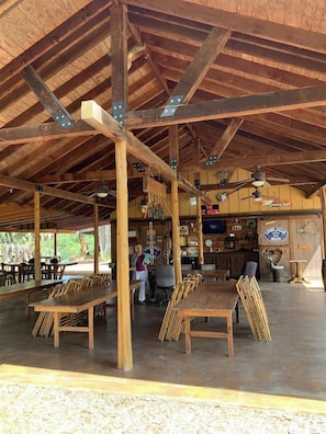 A covered outdoor dining area with wooden tables and chairs, a bar area, and a sign for 'The Ice House.' - Madrona Casita | Romantic Stay w/ Hot Tub & Views (Pipe Creek)