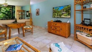 A room with a dining table, chairs, a cabinet, a television, and a large window with a view of greenery. - Affordable GEM/NewA/C, 1br/1ba w/Tropical Lanai, Steps to Ocean, LOCATION Unit 2 (Lahaina)