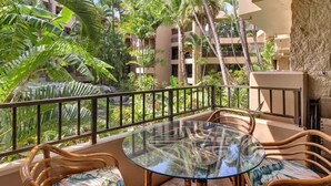 A balcony with a glass table and wicker chairs, surrounded by lush tropical plants and a view of a multi-story building. - Affordable GEM/NewA/C, 1br/1ba w/Tropical Lanai, Steps to Ocean, LOCATION Unit 2 (Lahaina)