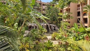 A lush garden with a waterfall, palm trees, and a building with balconies. - Affordable GEM/NewA/C, 1br/1ba w/Tropical Lanai, Steps to Ocean, LOCATION Unit 2 (Lahaina)