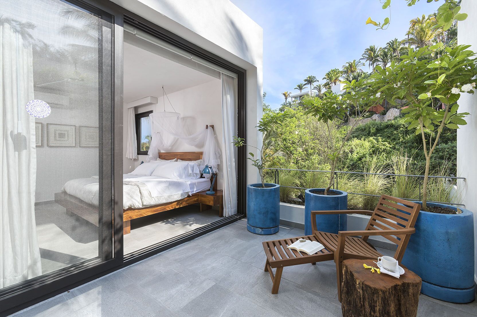 A modern bedroom with a large sliding glass door leading to a balcony with a wooden bench, a small table, and potted plants.