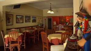 A dining area with wooden tables and chairs, a kitchen area in the background, and wall-mounted artwork.