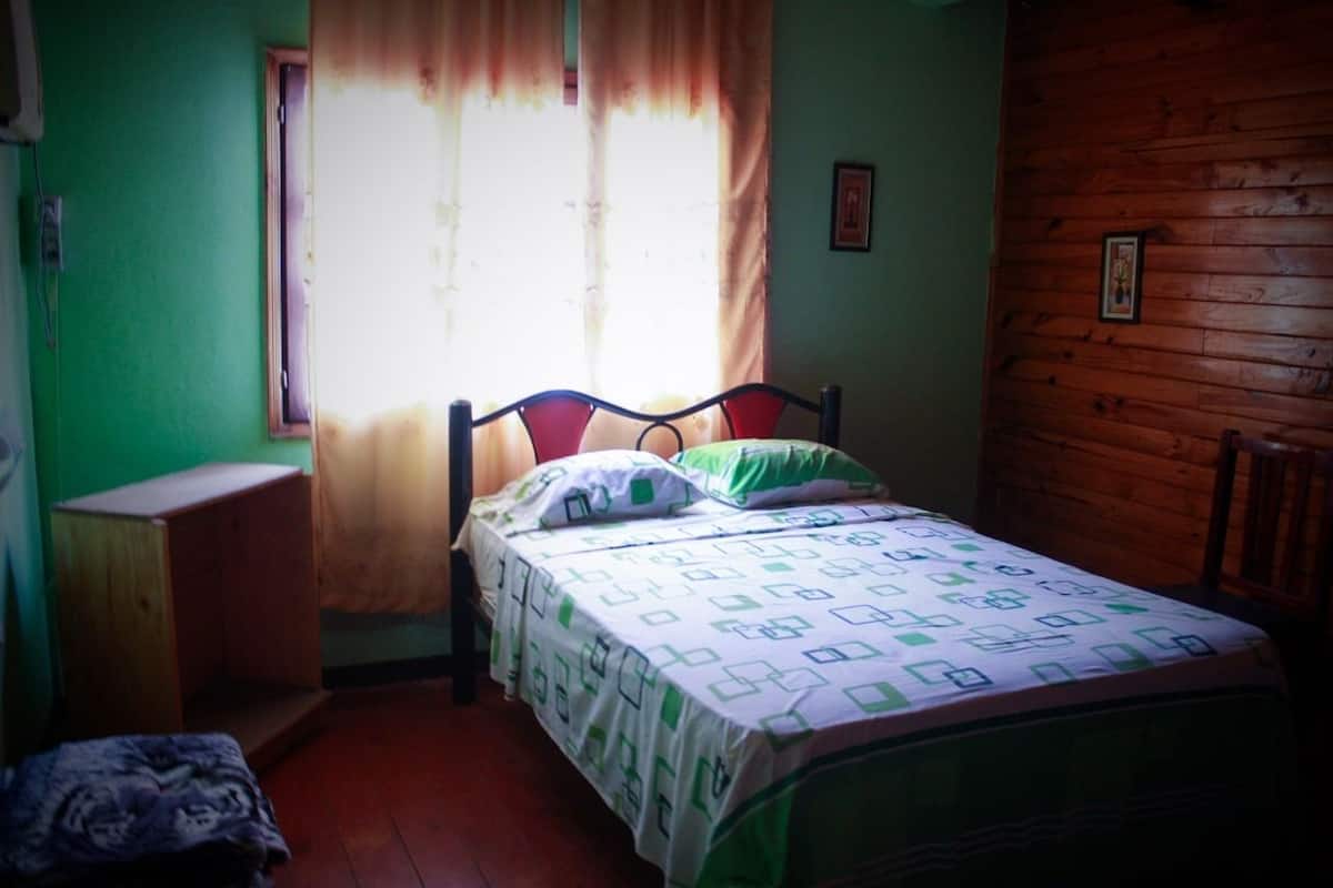 Quarto pequeno com uma cama de solteiro, cabeceira de madeira, janela com cortinas e parede verde.