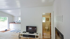 Una sala moderna con un televisor de pantalla plana sobre un mueble blanco, una chimenea y vistas a la cocina y el baño a través de una puerta abierta.