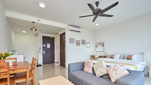 Una habitación de hotel moderna con una cama, un sofá gris, una mesa de comedor de madera con sillas, un ventilador de techo y una kitchenette.