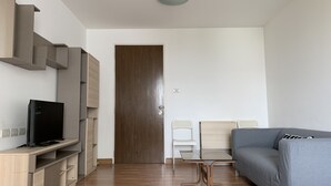 A modern living room with a wooden floor, a grey sofa, a TV on a wooden stand, and a door in the background. - Quiet & Secluded Urban Sanctuary for 2 (Bangkok)