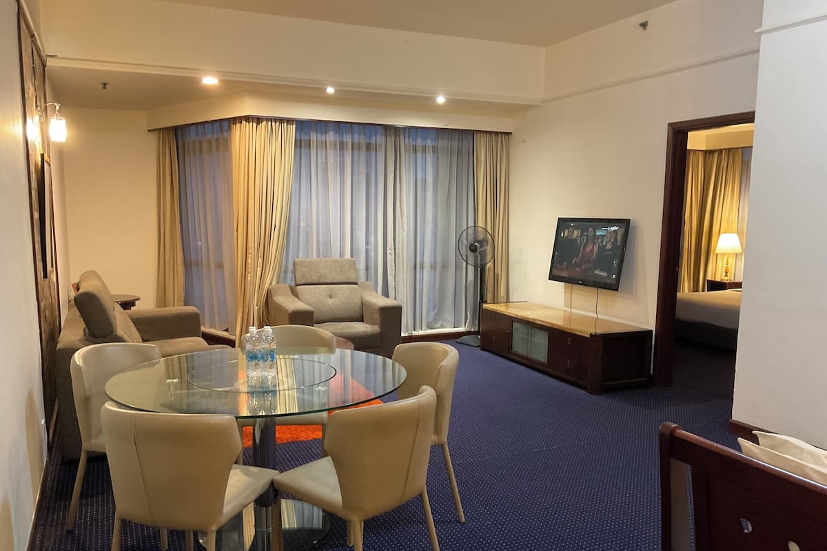 A hotel room with a glass-top dining table, two armchairs, a sofa, a TV on a stand, and a large window with curtains.