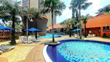 A swimming pool area with lounge chairs and umbrellas, surrounded by palm trees and a building with a curved facade.