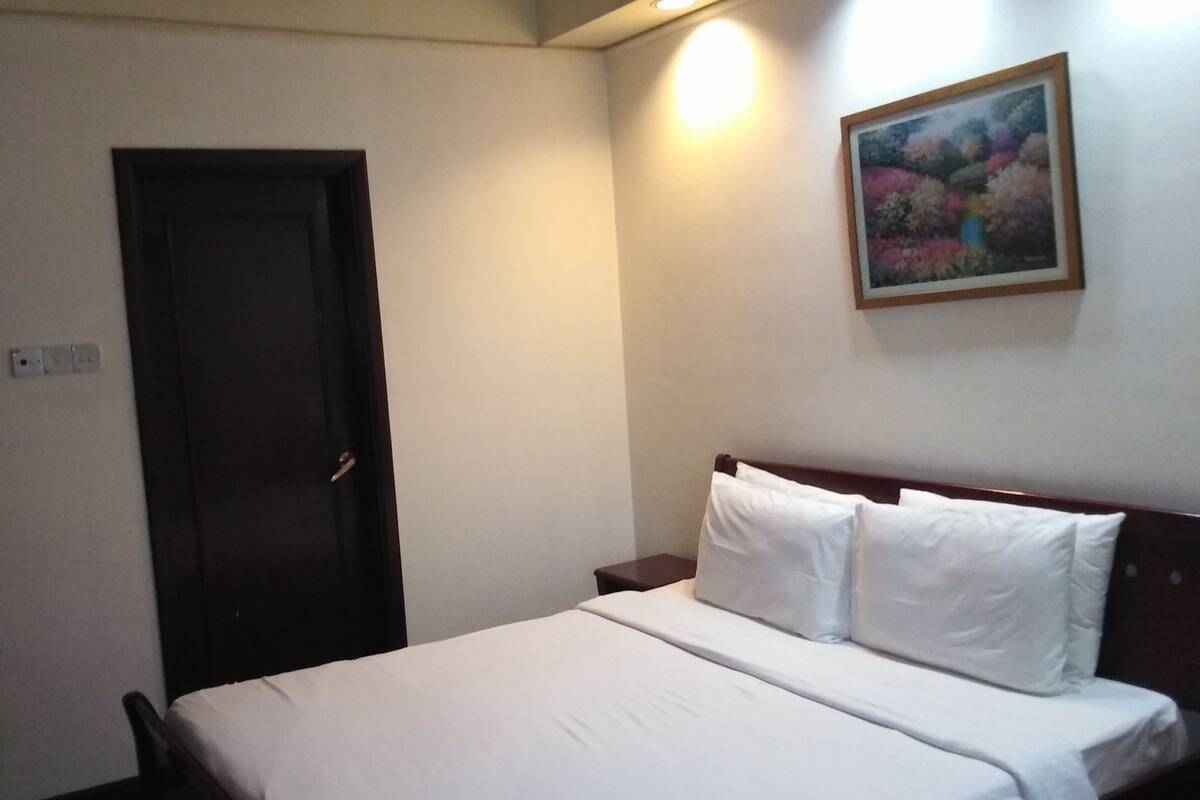 A hotel room with a bed, a door, a nightstand, a wall-mounted painting, and a ceiling light.