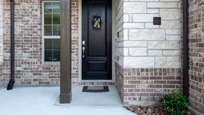 Une porte noire ornée d’un panneau de verre décoratif, une façade en briques et une petite plante en pot près de l’entrée.