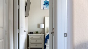 Un couloir étroit mène à une chambre où se trouvent une commode blanche, une lampe et une œuvre d’art encadrée accrochée au mur.