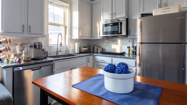 Une cuisine moderne avec des meubles blancs, des appareils en acier inoxydable et une table en bois surmontée d’un set de table bleu.
