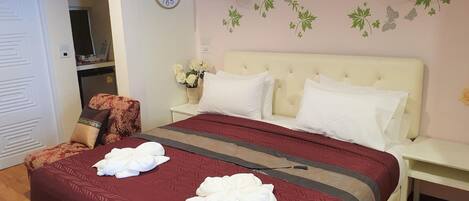 Habitación de hotel con una cama grande, dos almohadas blancas, una colcha marrón y roja, un reloj en la pared, una lámpara de pared y una pequeña mesa con un jarrón de flores.