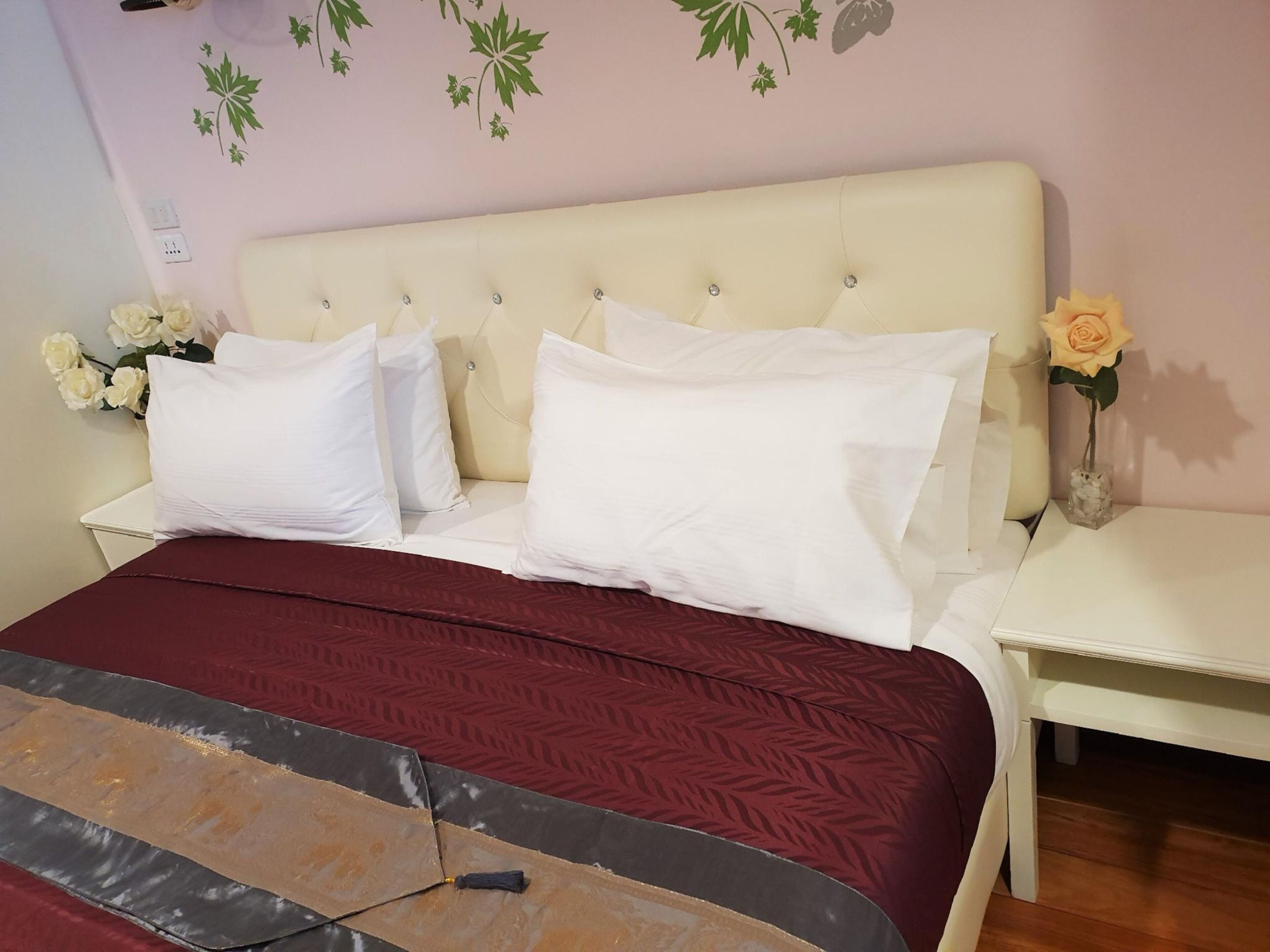 Una cama bien hecha con ropa blanca, un edredón rojo y un cabecero decorativo. Hay mesitas de noche blancas con un jarrón de flores en cada una.