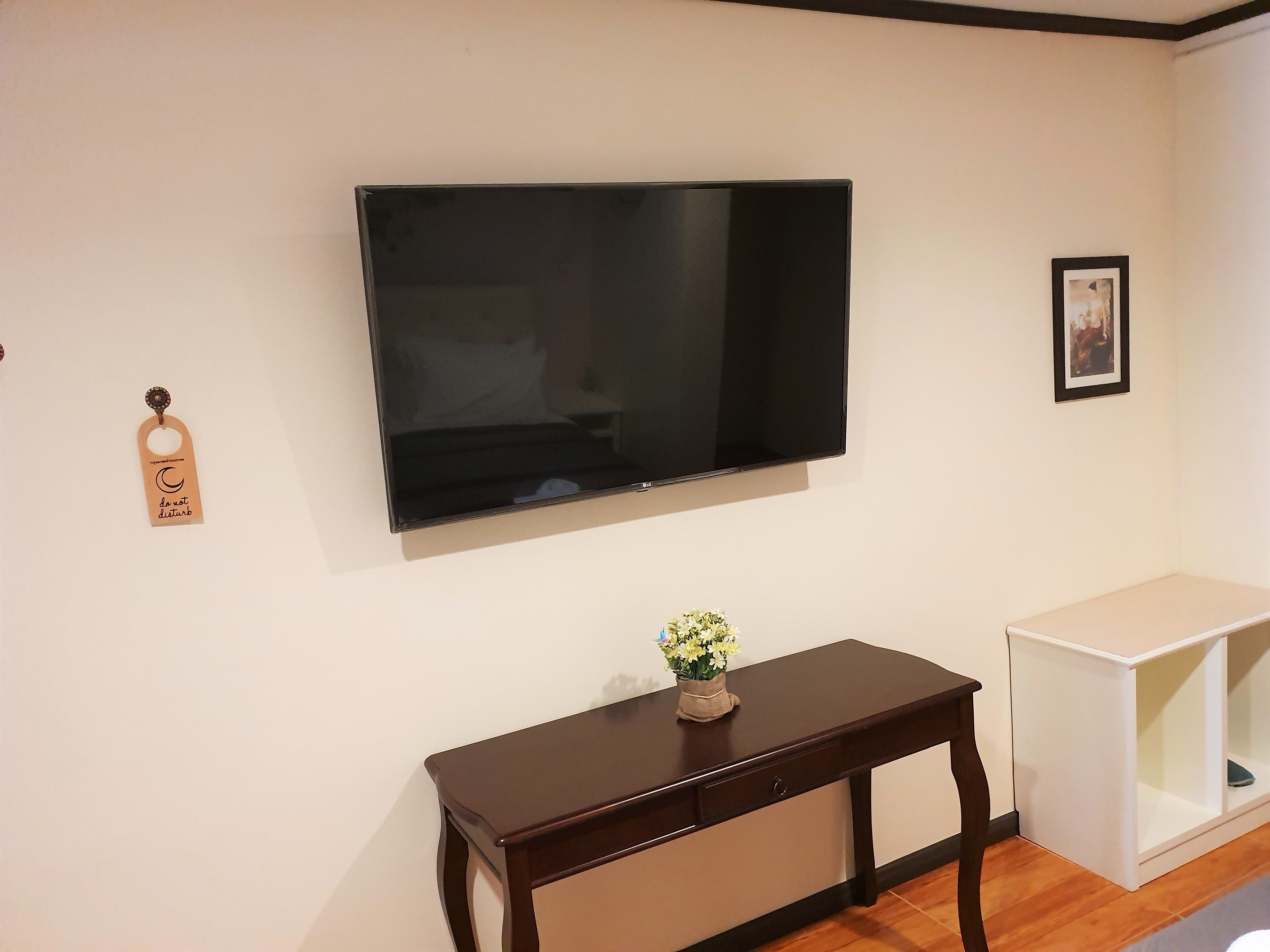 A living room with a flat-screen TV mounted on the wall, a wooden side table with a vase of flowers, and a white cabinet.