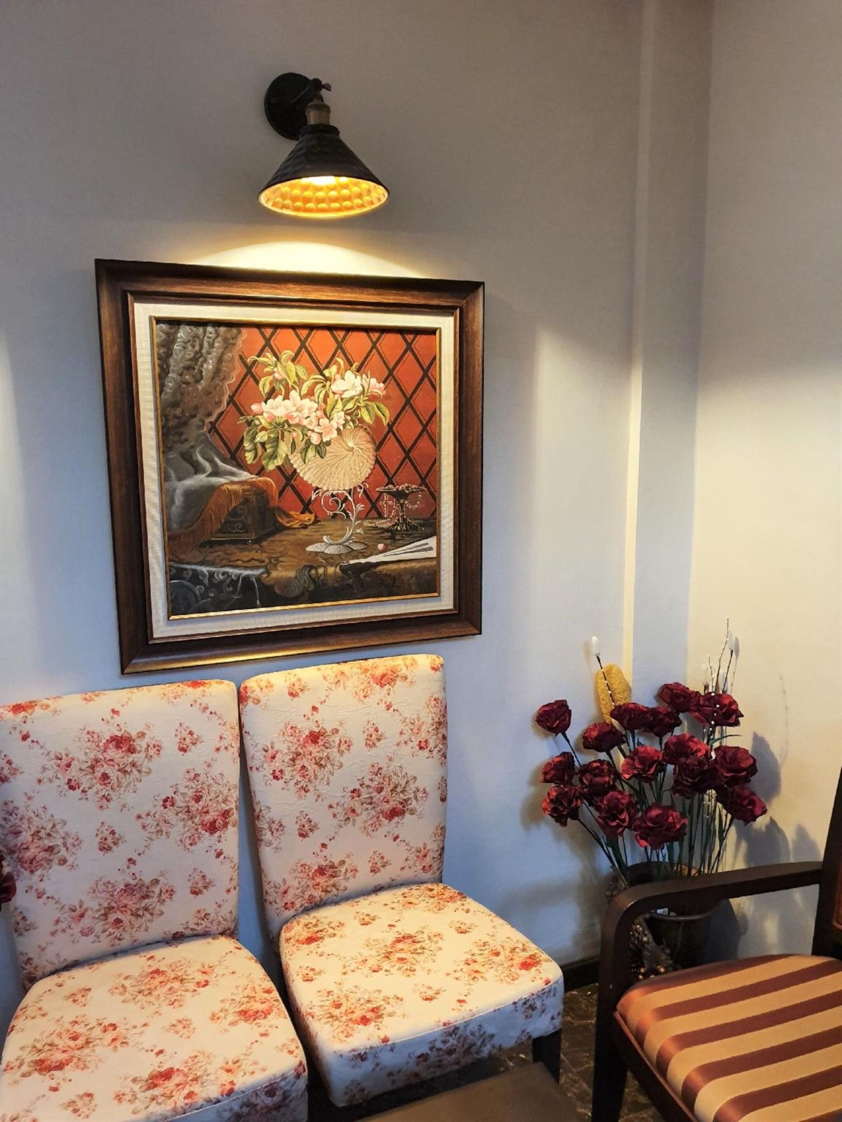 A room with two floral-patterned chairs, a framed painting above them, and a vase with red flowers on a table.