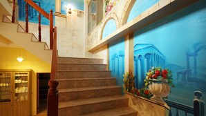 Una gran escalera con escalones de mármol y balaustres de madera, que conduce a un rellano con un mural decorativo de arquitectura clásica.