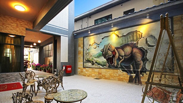Un espacio al aire libre con sillas y mesas de metal decorativo, un mural de un dinosaurio en la pared de un edificio y un extintor.