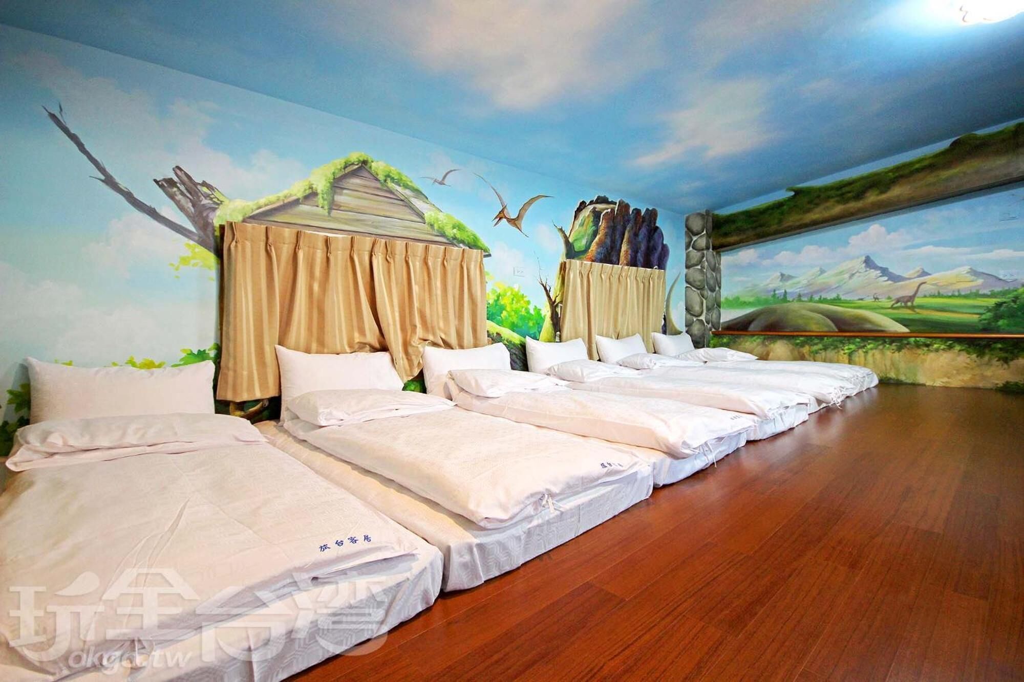 Une chambre d’hôtel avec quatre lits disposés en rangée, chacun étant surmonté d’un baldaquin. Un mur de la chambre est décoré d’une fresque représentant un paysage, situé derrière les lits.