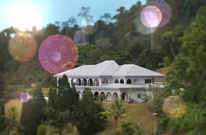 A large, white building with multiple arches and a sloped roof, surrounded by lush greenery.
