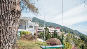 A view of a building with a large tree in the foreground, a swing set, and a mountainous landscape in the background. - Cozy Villa Genting Highlands (Genting Highlands)