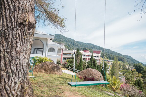 A view of a building with a large tree in the foreground, a swing set, and a mountainous landscape in the background.