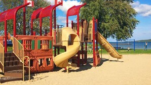 Une aire de jeux avec un toboggan et des structures d’escalade, située sur une zone sablonneuse, avec un lac en arrière-plan.