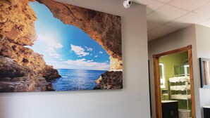 Un couloir orné d’un grand tableau représentant un paysage côtier, une porte en bois donnant sur une pièce dont un mur est recouvert de carreaux verts, et un lavabo blanc avec un miroir.