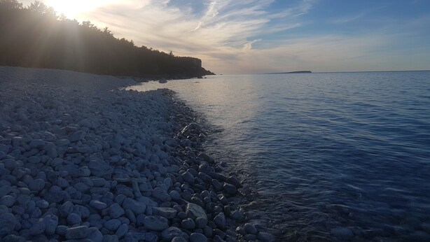 Une côte rocheuse bordée d’une eau calme, un ciel dégagé et une terre lointaine à l’horizon.