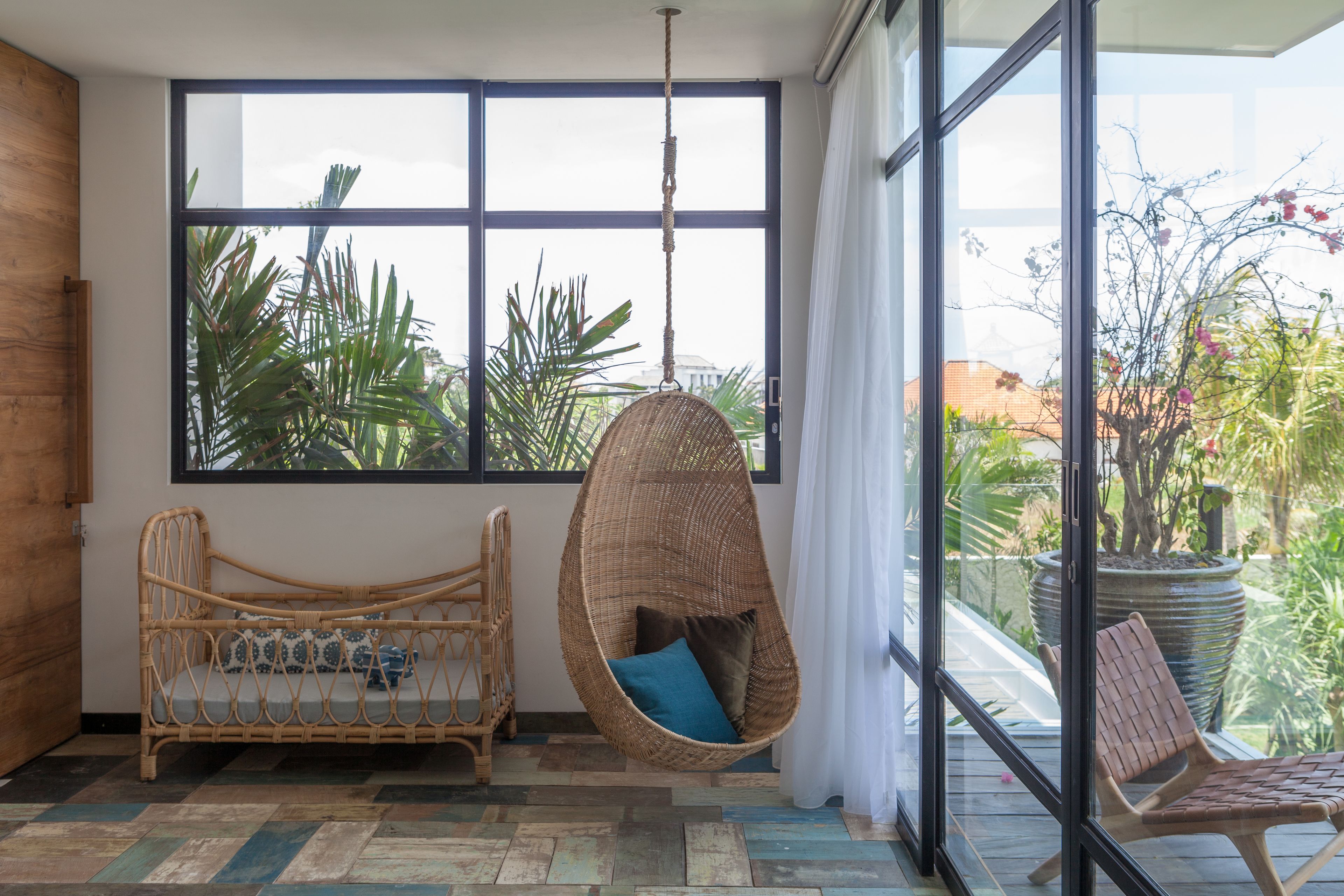 A room with a large window, a hanging chair, a wicker sofa, and a view of greenery outside.