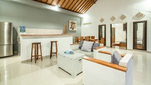 Cozinha moderna com geladeira de aço inoxidável, dispenser de água e área de bar com banquetas. Há um espaço de estar com móveis de vime e um espelho grande.