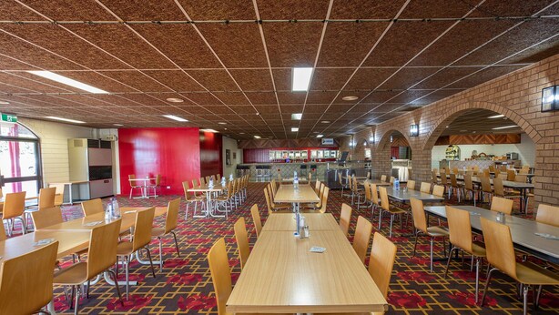 A spacious cafeteria with multiple tables and chairs, a patterned carpet, and a brick wall with a kitchen area in the background. - Leichhardt Hotel Motel (Cloncurry)