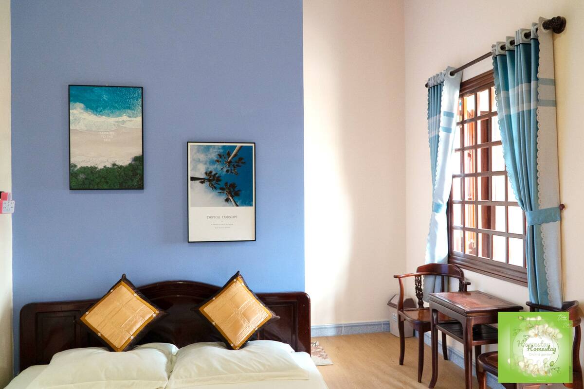 Une chambre à coucher avec un lit, deux oreillers, une table de chevet, une fenêtre avec des rideaux et deux tableaux encadrés accrochés au mur.