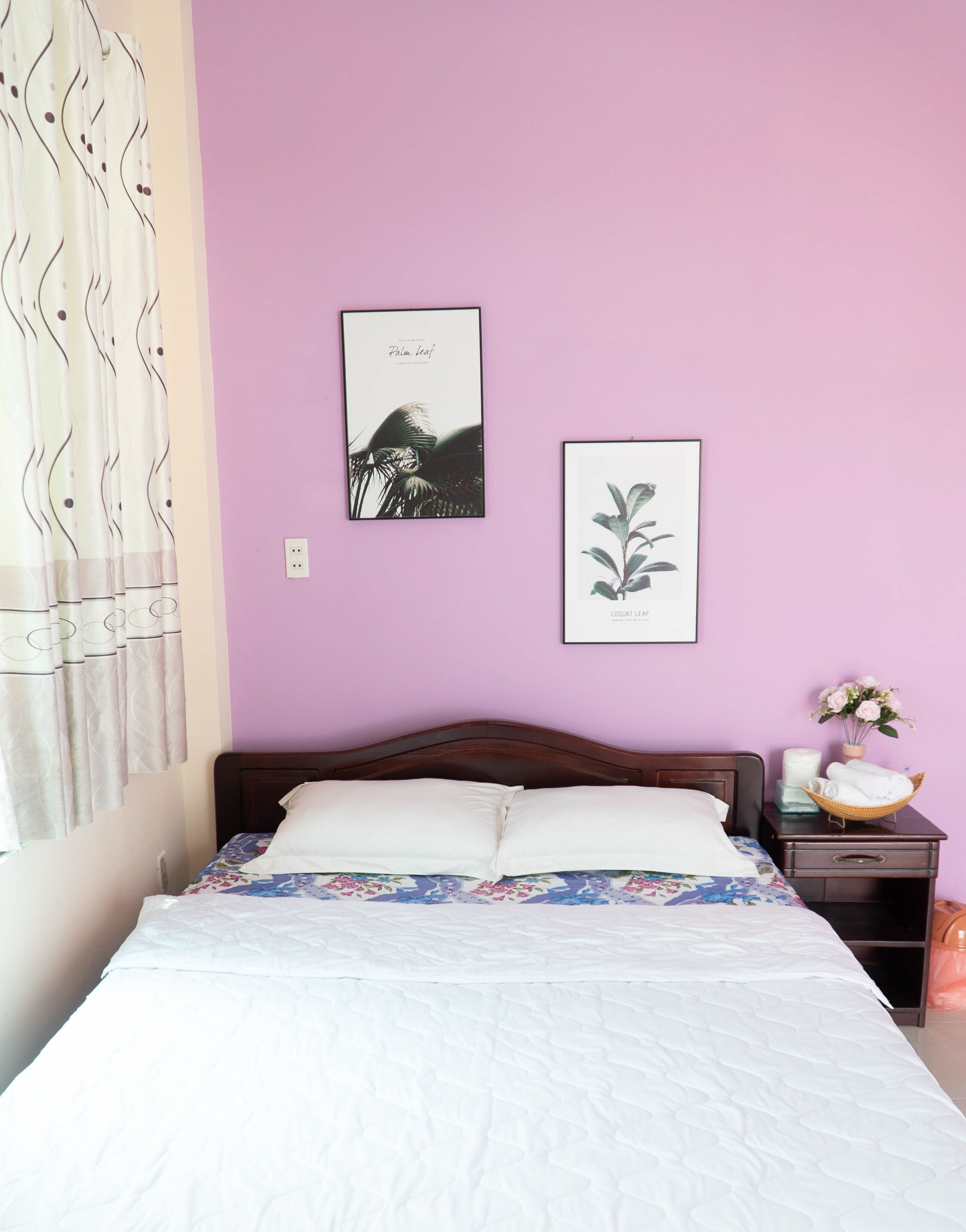 A bedroom with a bed, a nightstand, a wall-mounted power outlet, and two framed botanical prints.