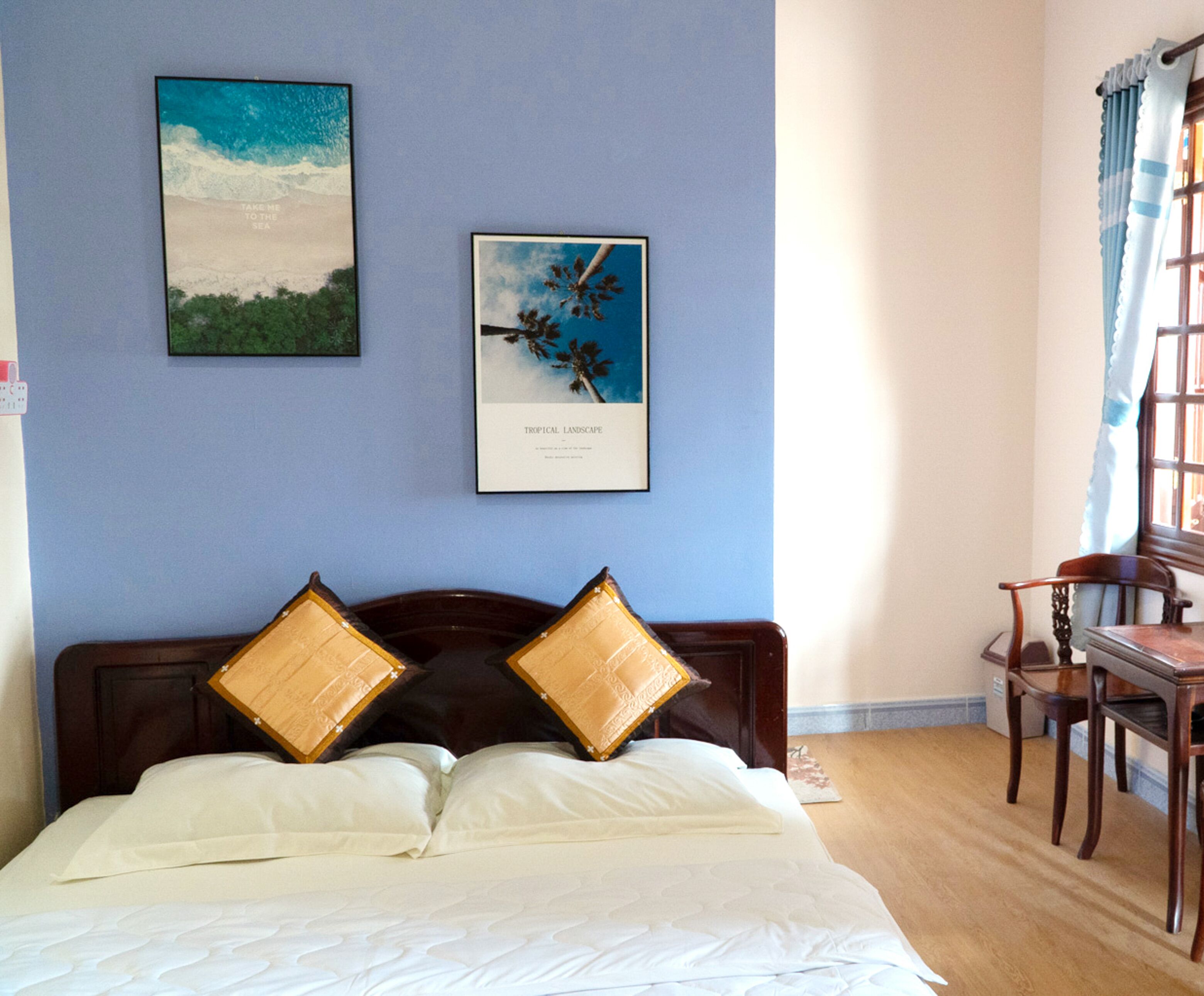 A hotel room with a bed, two framed pictures on the wall, a wooden chair, and a window with curtains.