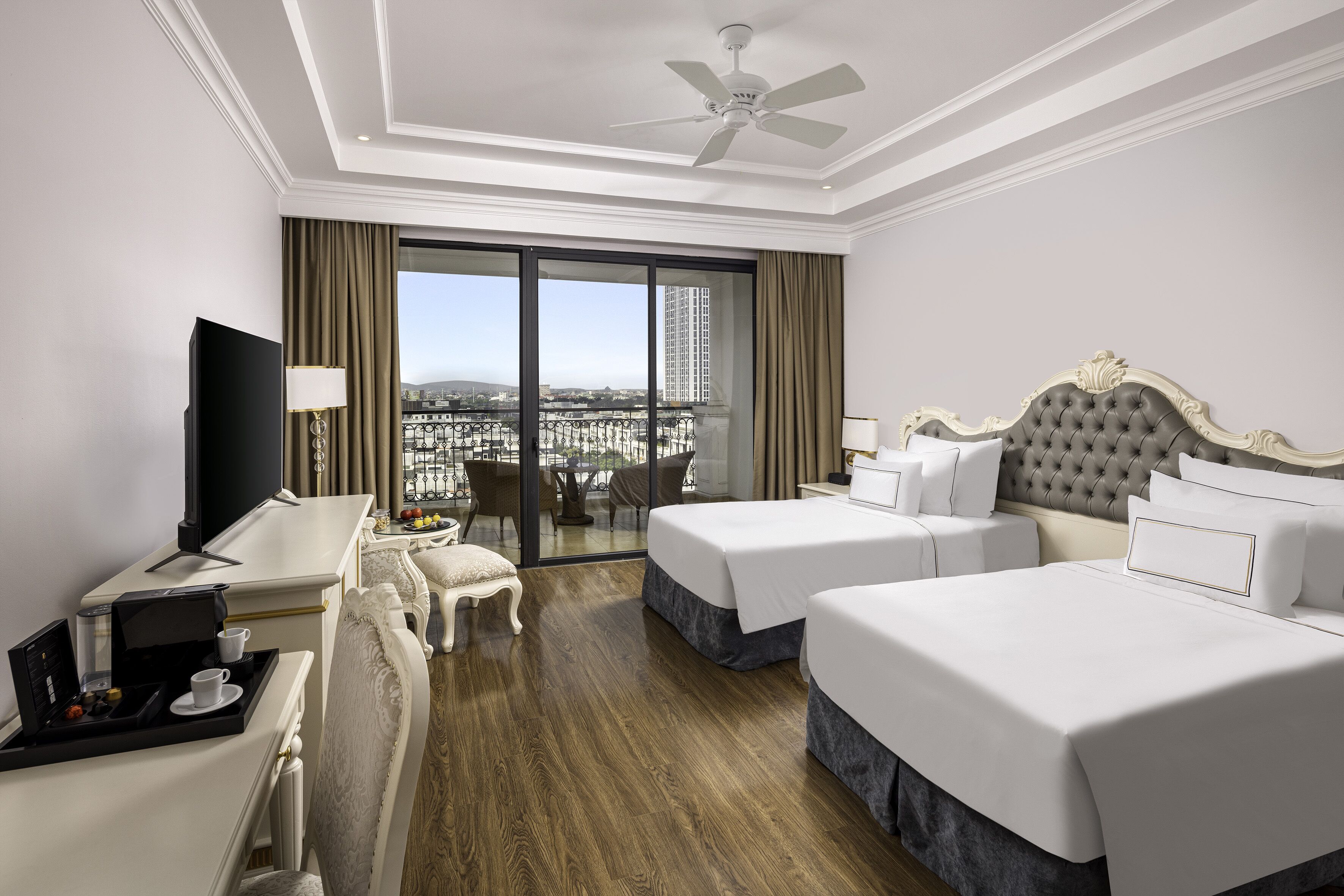 Habitación de hotel con dos camas, un escritorio con cafetera, televisor y balcón con vista a la ciudad.