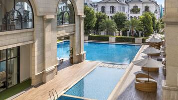 A pool area with a clear view of the surrounding architecture, including a large building with arched windows and a poolside lounge with umbrellas.