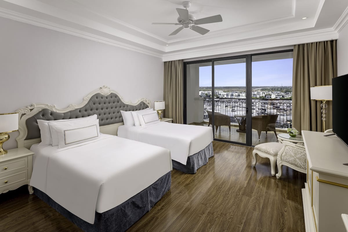 A hotel room with two beds, a balcony with a city view, a desk with a chair, and a ceiling fan.